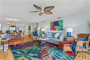 A spacious living area with a ceiling fan, a colorful abstract painting, and a dining area with wicker furniture. - Isle Be Back, Oceanfront Escape sleeps 4 (St Simons Island)