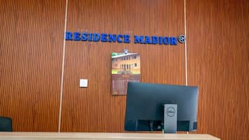 Un mostrador de recepción con un panel de pared de madera, un cartel que dice 'RESIDENCE MADIOR', un monitor de computadora Dell y un calendario con el año 2025.