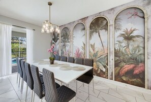 Zona de menjador amb taula blanca, cadirees amb seients negres i un mural tropical a la paret.