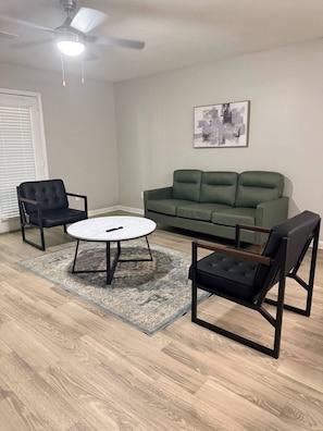 Ein Wohnzimmer mit Deckenventilator, einem grünen Sofa, zwei Sesseln und einem runden Couchtisch.