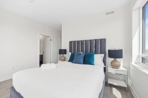 A modern hotel room with a large bed, two bedside tables, a window, and a door leading to another room. - Luxury 3BR Penthouse • Central Location + City Views + Parking (Toronto)