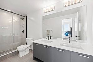 Et moderne badeværelse med en glasbrusekabine, et hvidt toilet og et håndvaskarrangement med dobbeltvask og et stort spejl.