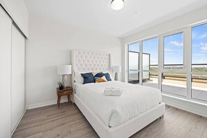 A hotel room with a large bed, a nightstand, a lamp, and a view of the city through large windows. - Luxury 3BR Penthouse • Central Location + City Views + Parking (Toronto)