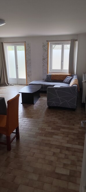 A modern living room with a sofa, ottoman, and a chair. There are large windows with curtains and a radiator on the wall. - Spacious House in the Peaceful Countryside (Val-Sonnette)