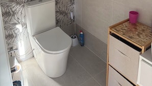 Ein Badezimmer mit Toilette, Bidet, einem kleinen Holztisch und einer rosa Tasse, sowie einer Wand mit Blätermuster.