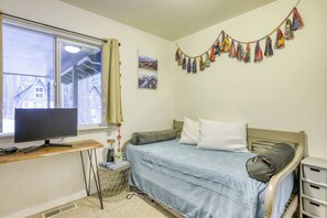 Une chambre avec un lit, un bureau doté d’un écran d’ordinateur, une fenêtre donnant sur une maison et un mur orné de tableaux et d’une guirlande en tissu.