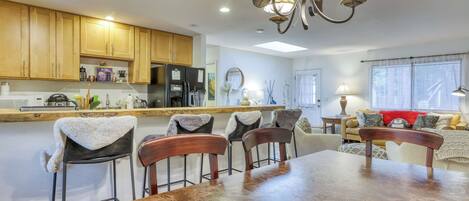 A modern kitchen with wooden cabinets, a dining table with chairs, a chandelier, and a living area with a sofa and a window.