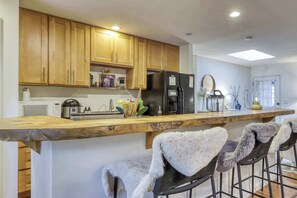 A kitchen with wooden cabinets, a refrigerator, a coffee maker, and bar stools with fur throw blankets. - Gardner Mountain Hideaway (South Lake Tahoe)