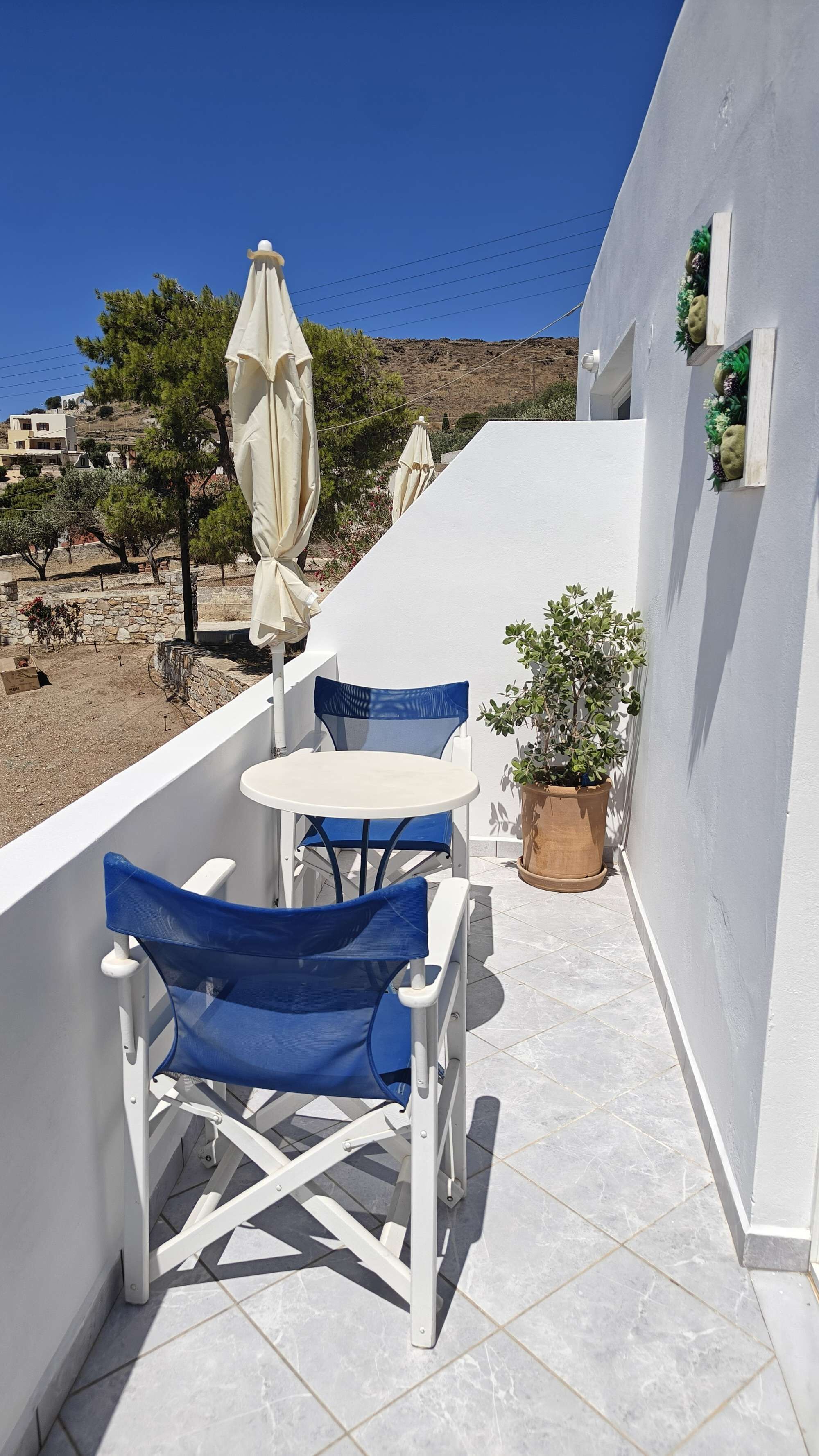 A small outdoor balcony with a white table and blue chairs, a potted plant, and a view of the landscape.