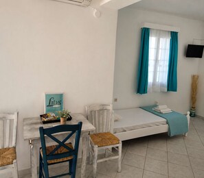 A small, clean room with a bed, a table with chairs, a TV, and a window with curtains. - Sea View Studios in Syros – Litus Studios (Syros)