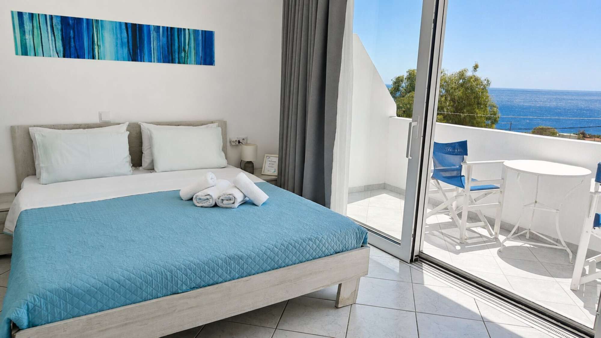 A hotel room with a bed, a view of the sea, and a balcony with a table and chairs.