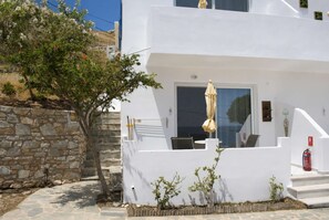 A white building with a stone wall, a door, and a small garden area with plants and a tree. - Sea View Studios in Syros – Litus Studios (Syros)