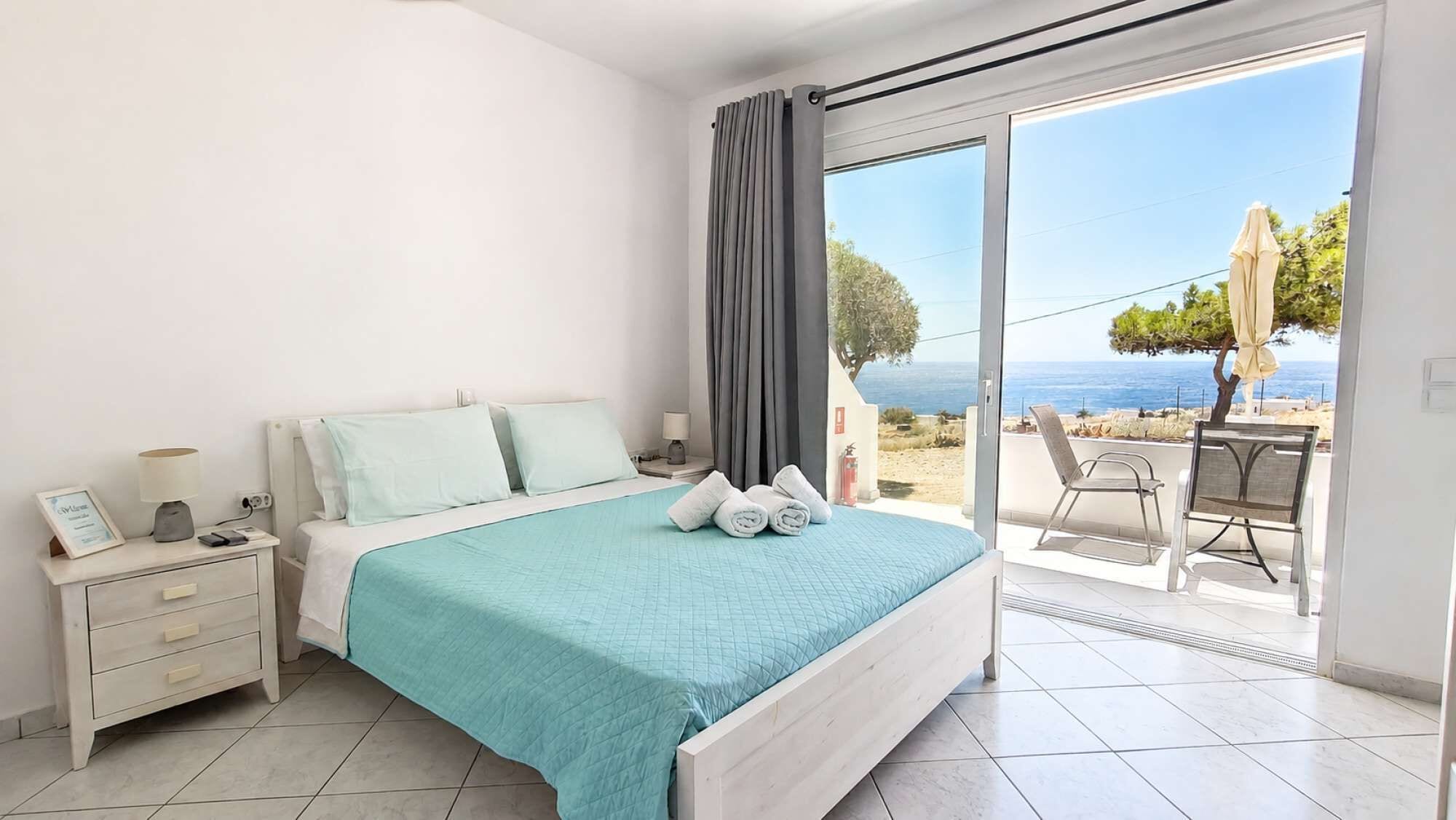 A bedroom with a bed, bedside tables, a nightstand, a view of the sea, and a balcony with chairs.