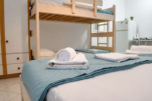 A bunk bed room with a blue quilted bedspread, a wooden dresser with drawers, and a white wall with a window. - Sea View Studios in Syros – Litus Studios (Syros)
