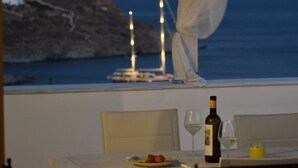 Mesa de jantar com garrafa de vinho, copos e prato de comida, com vista para um litoral rochoso com um veleiro e uma lua no céu.