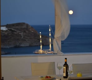 A dining table with a bottle of wine, glasses, and a plate of food, overlooking a rocky coastline with a sailboat and a moon in the sky. - Sea View Studios in Syros – Litus Studios (Syros)