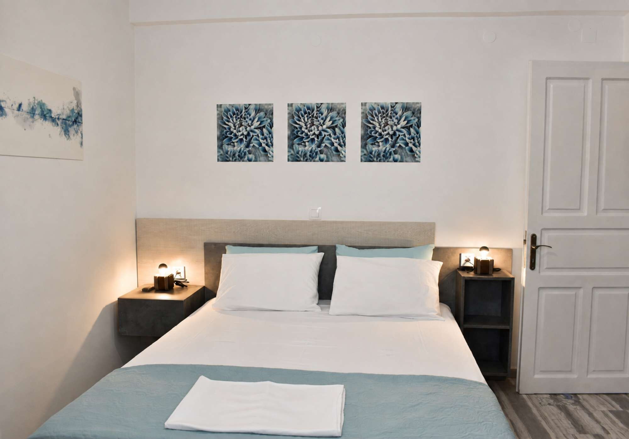 A hotel room with a bed, two bedside tables with lamps, a door, and three wall-mounted artworks.