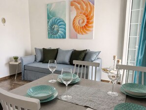 A dining area with a table set for two, a sofa, and two wall-mounted artworks. - Sea View Studios in Syros – Litus Studios (Syros)