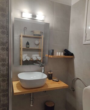 A bathroom with a large white sink, a mirror, and a shelf with towels and toiletries. - Sea View Studios in Syros – Litus Studios (Syros)