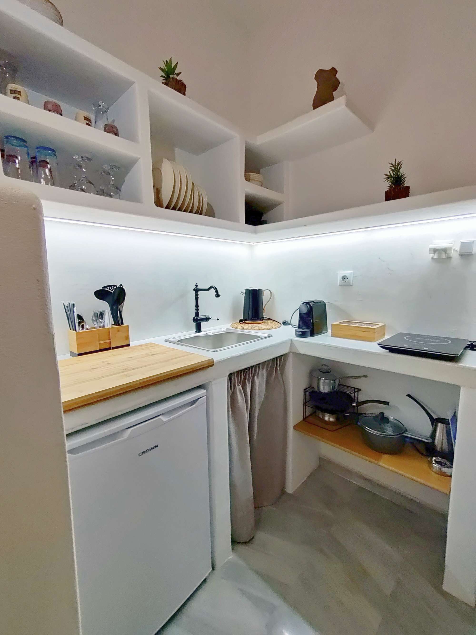 A compact kitchen with a built-in dishwasher, a sink, and a small refrigerator. There are shelves above the sink holding glassware and a potted plant.