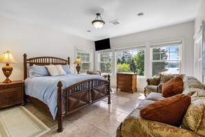 Une chambre avec un lit en bois, une table de chevet avec une lampe, une commode, un canapé avec des coussins et de grandes fenêtres donnant sur un paysage verdoyant et un lac.
