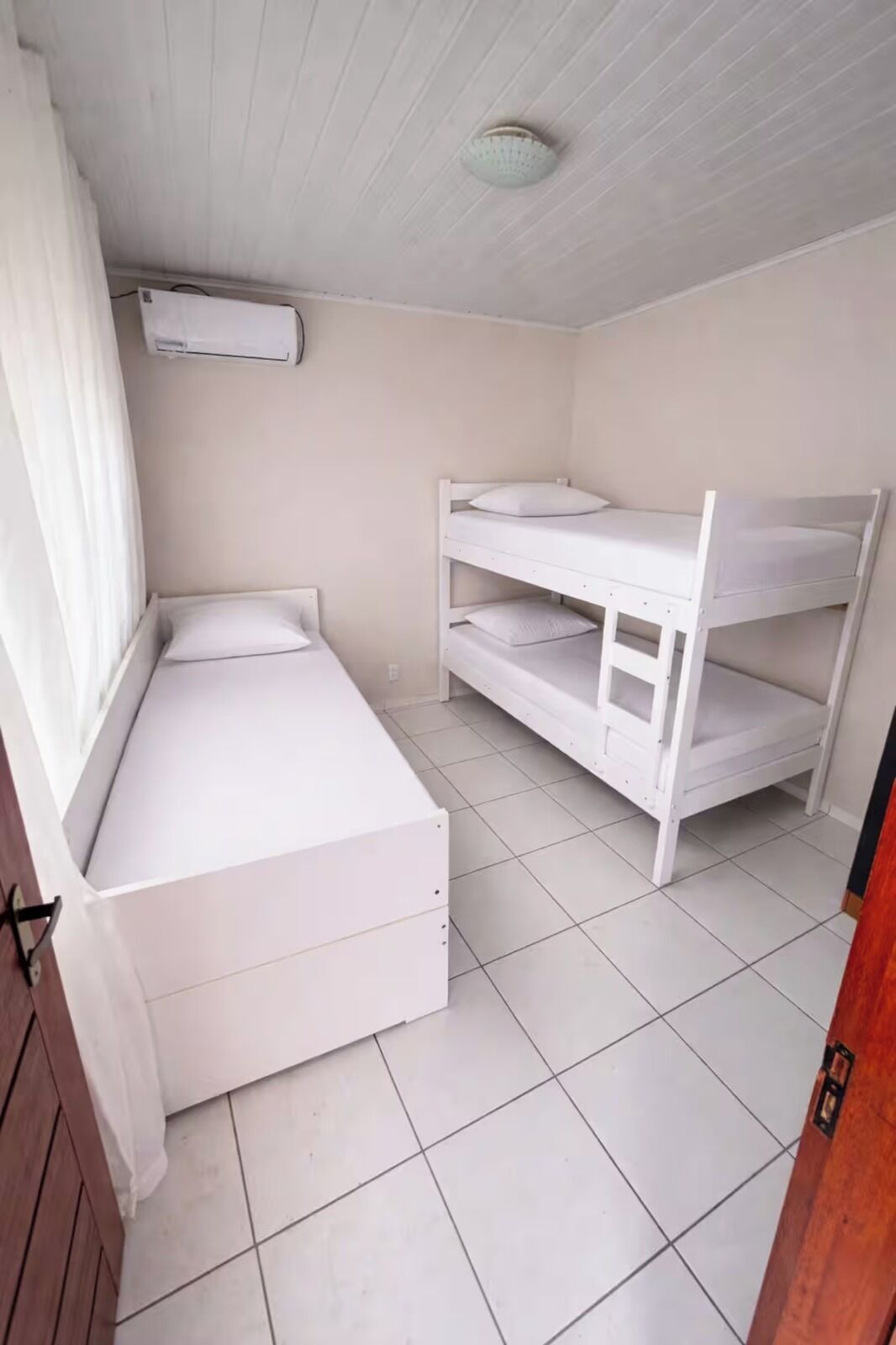 Quarto com beliche, uma cama de solteiro, janela com cortinas e ventilador de teto.