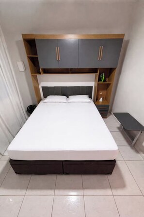 Un llit amb capçalera gris, una prestatgeria de fusta a sobre i una petita taula de nit amb una làmpada.