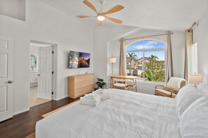 Une chambre avec un grand lit, un bureau, une chaise, une télévision et une fenêtre donnant sur des maisons et des palmiers.