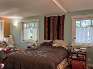 A bedroom with a large bed, two bedside tables, a chair, a bookshelf, and two windows with curtains. - artist's home (Boulder)