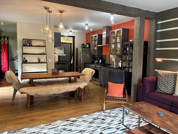 A modern kitchen with stainless steel appliances, a dining area with a wooden table and chairs, and a living room with a sofa and patterned rug.