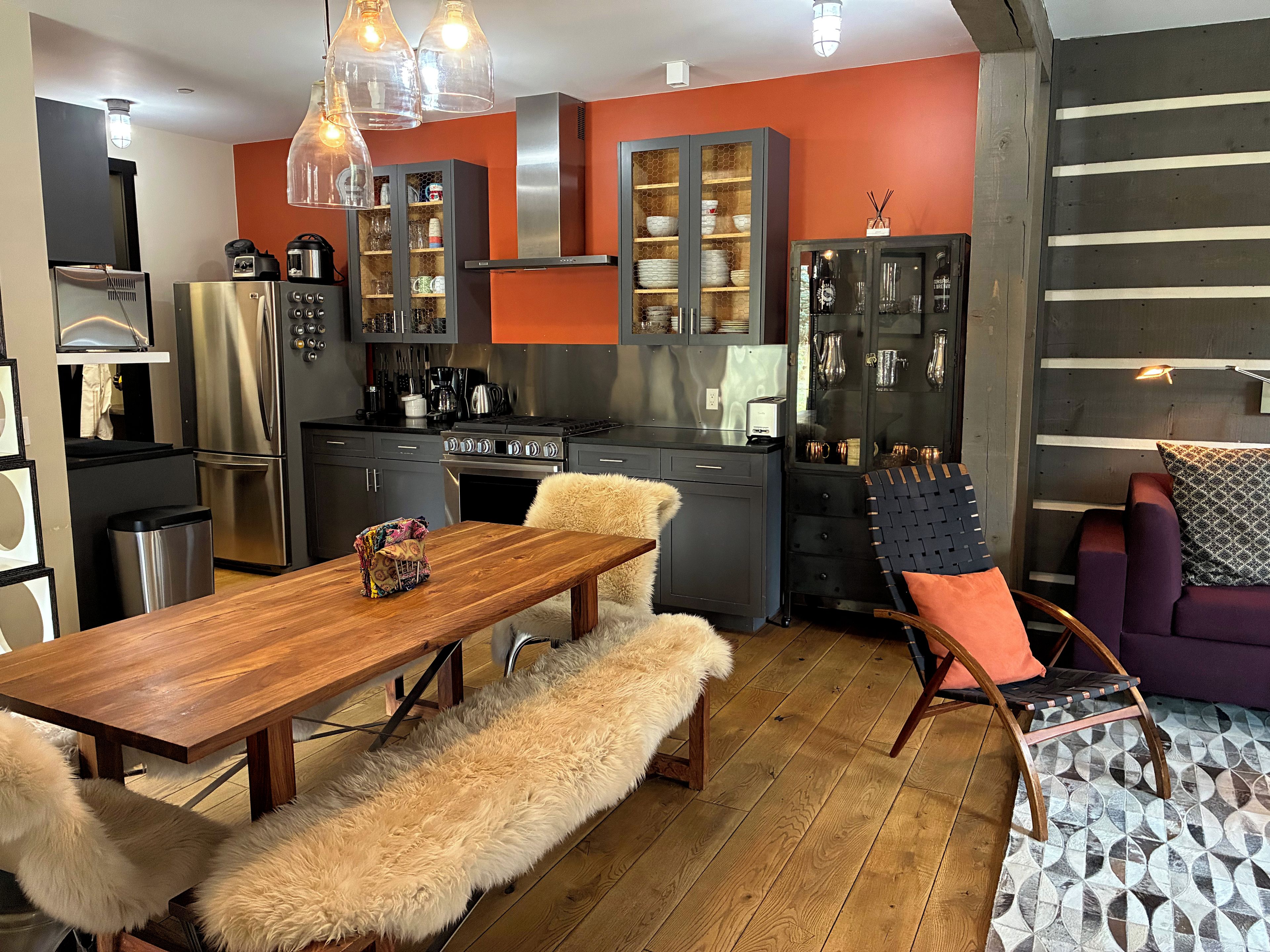 Una cocina moderna con electrodomésticos de acero inoxidable, un comedor con mesa de madera y manta de piel, y una sala de estar con sofá púrpura.