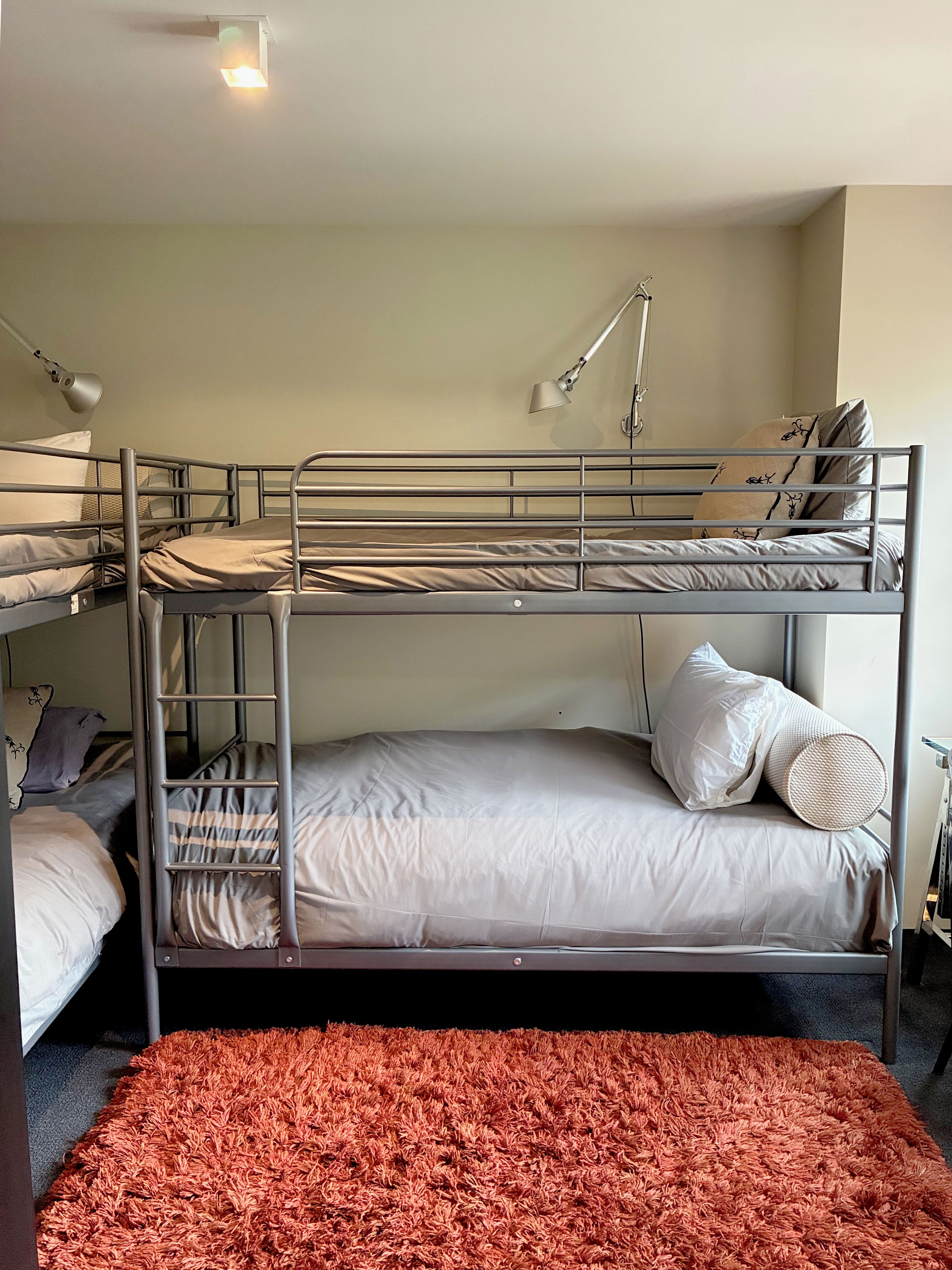 Habitación con literas, alfombra roja, dos lámparas de noche y una lámpara de pared.