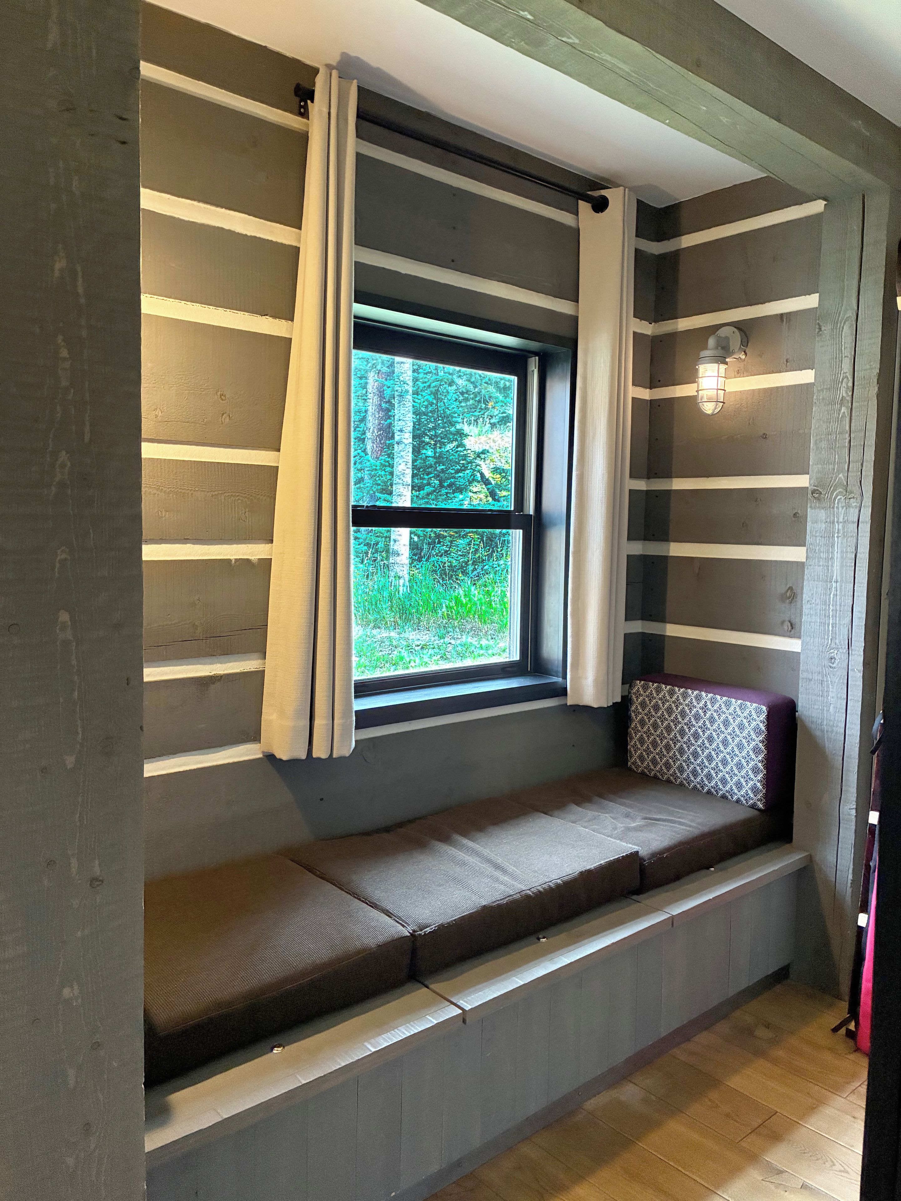 Ein Zimmer mit einer eingebauten Sitzbank unter einem Fenster mit Vorhängen, einer Wandleuchte und einem Holzboden.