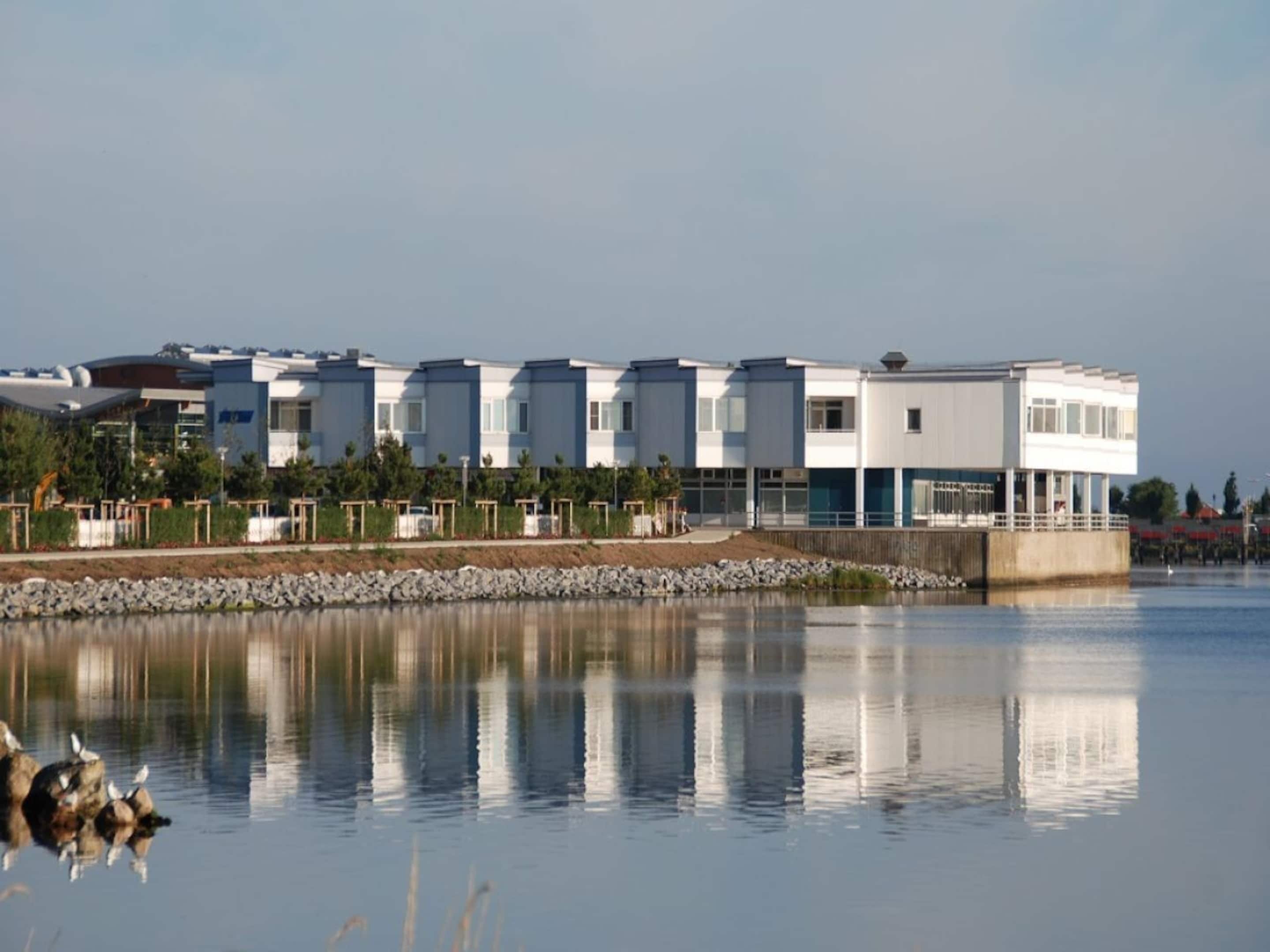 A modern building with a flat roof, large windows, and a reflective water body in front.