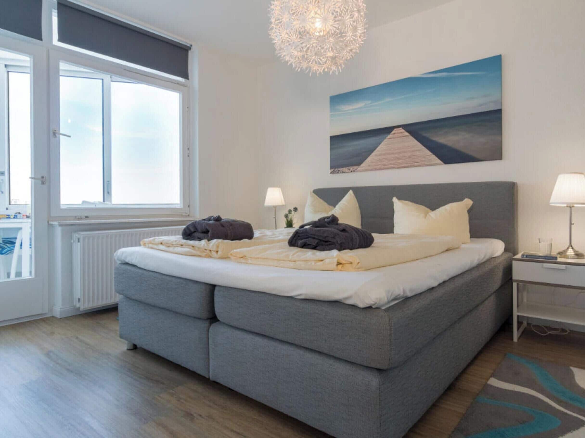 Quarto moderno com cama grande, luminárias de cabeceira, vista para o mar pela janela e um quadro na parede.