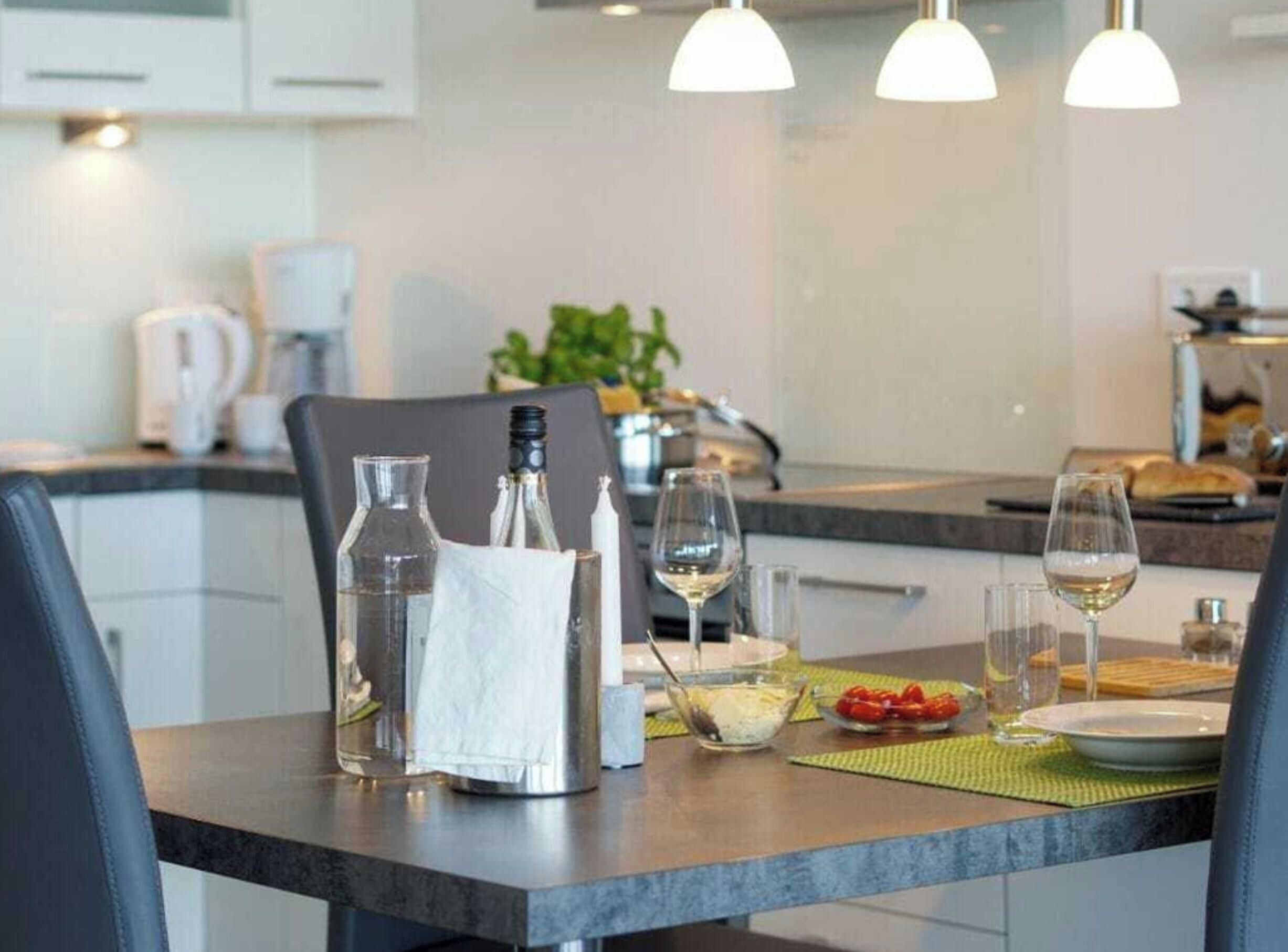 A modern kitchen with a dining table set for two, featuring a wine glass, a bowl of fruit, and a bottle of water.