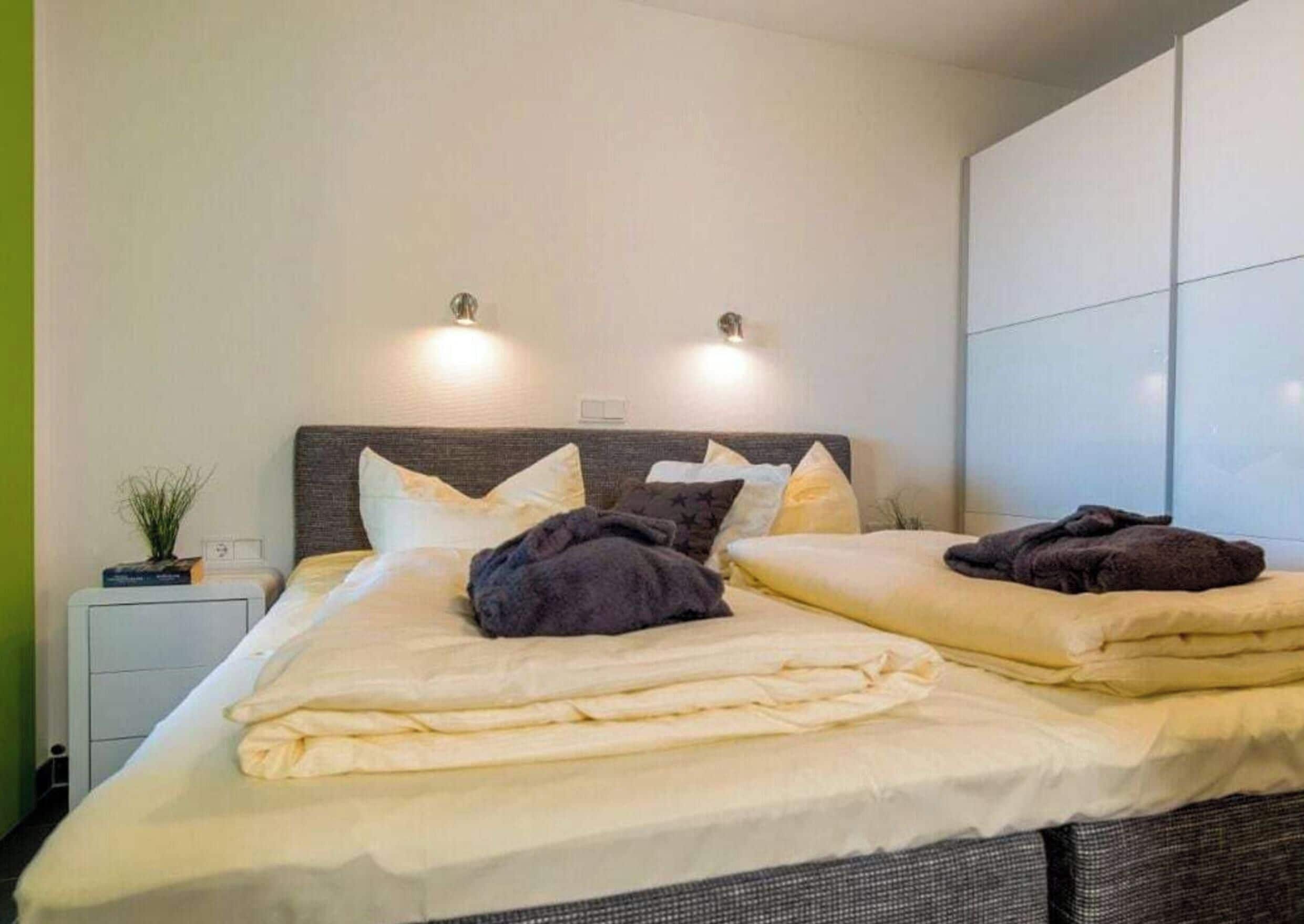 Uma cama de casal com cabeceira, duas luzes na parede e uma mesa de cabeceira com uma planta.