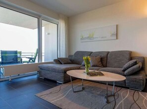 Ein modernes Wohnzimmer mit einem grauen Sofa, einem runden Couchtisch und Blick durch ein großes Fenster.