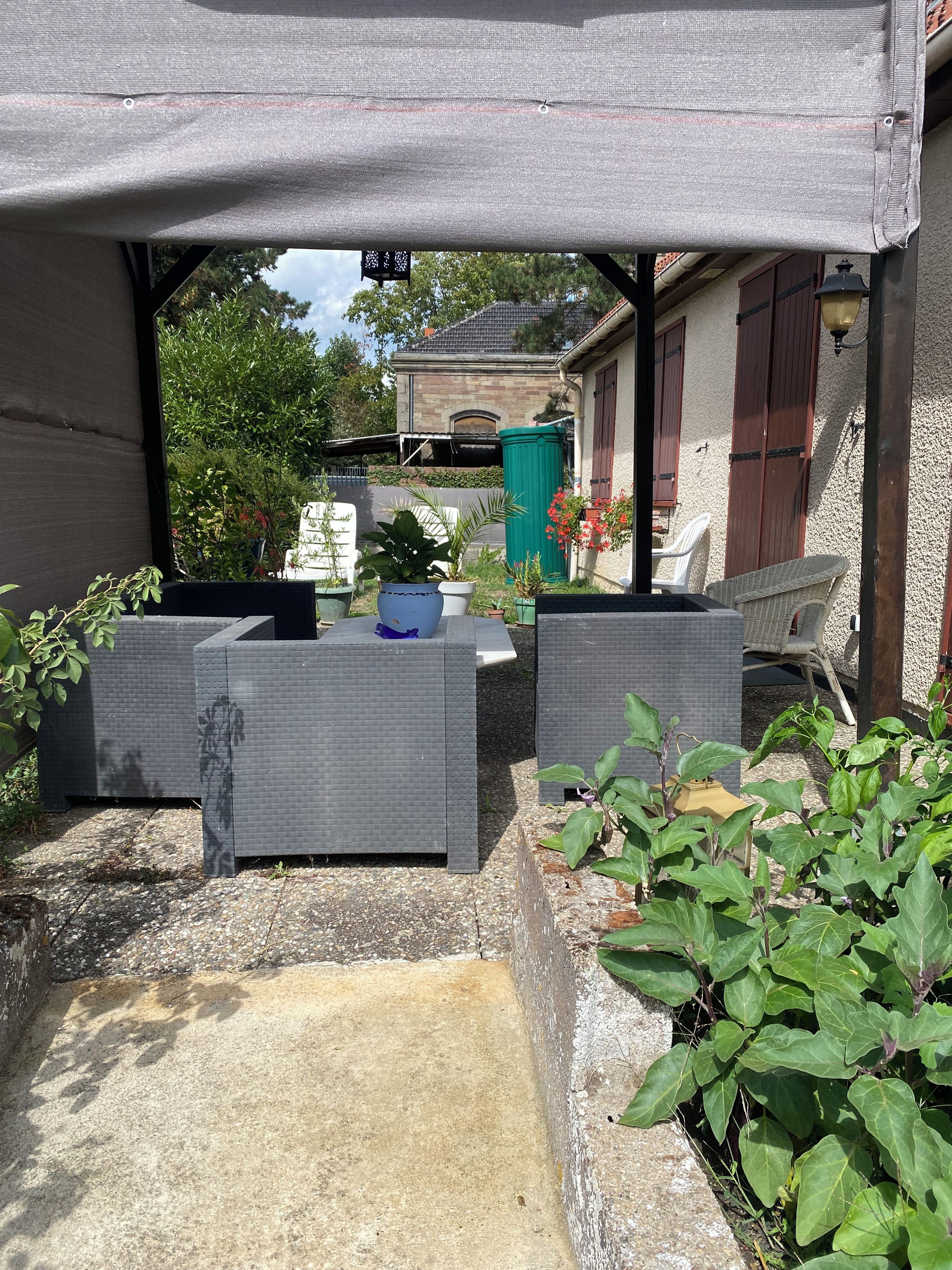 Un espacio al aire libre cubierto con mobiliario de mimbre, plantas en macetas y vista a una casa al fondo.