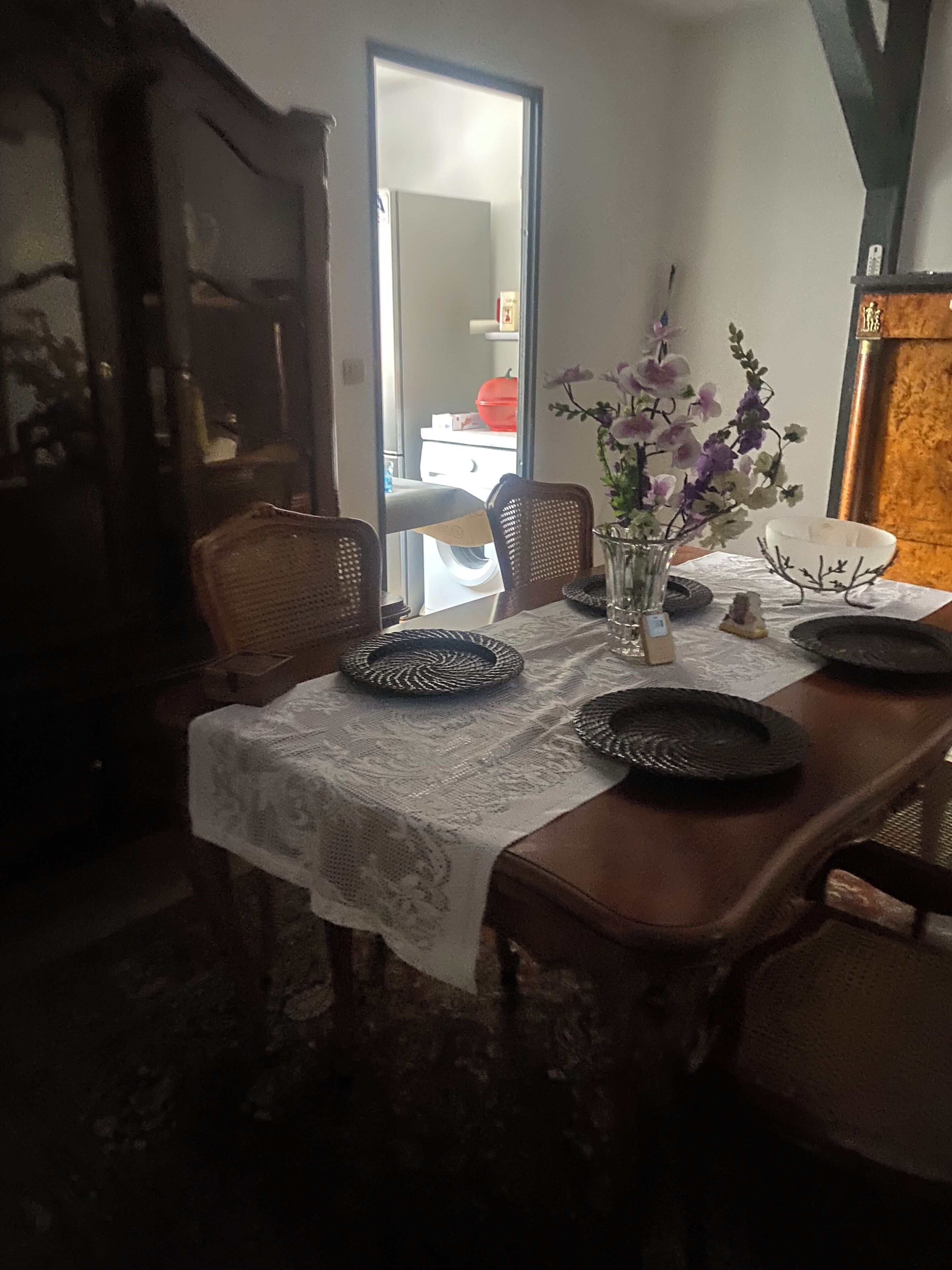 Una mesa de comedor con mantel de encaje, rodeada de sillas, un florero con flores y un tazón.