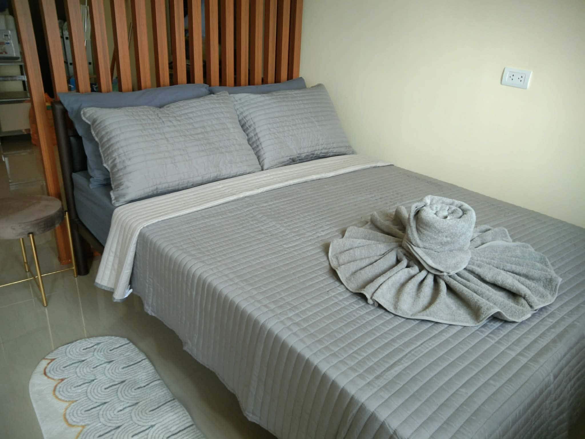 Un lit bien fait, avec une couette grise et des oreillers, un tapis rond à motifs ondulés et une tête de lit en bois.