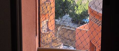 Une vue à travers une fenêtre donnant sur une clôture en treillis métallique, avec en arrière-plan un immeuble doté de nombreux balcons et un ciel dégagé.
