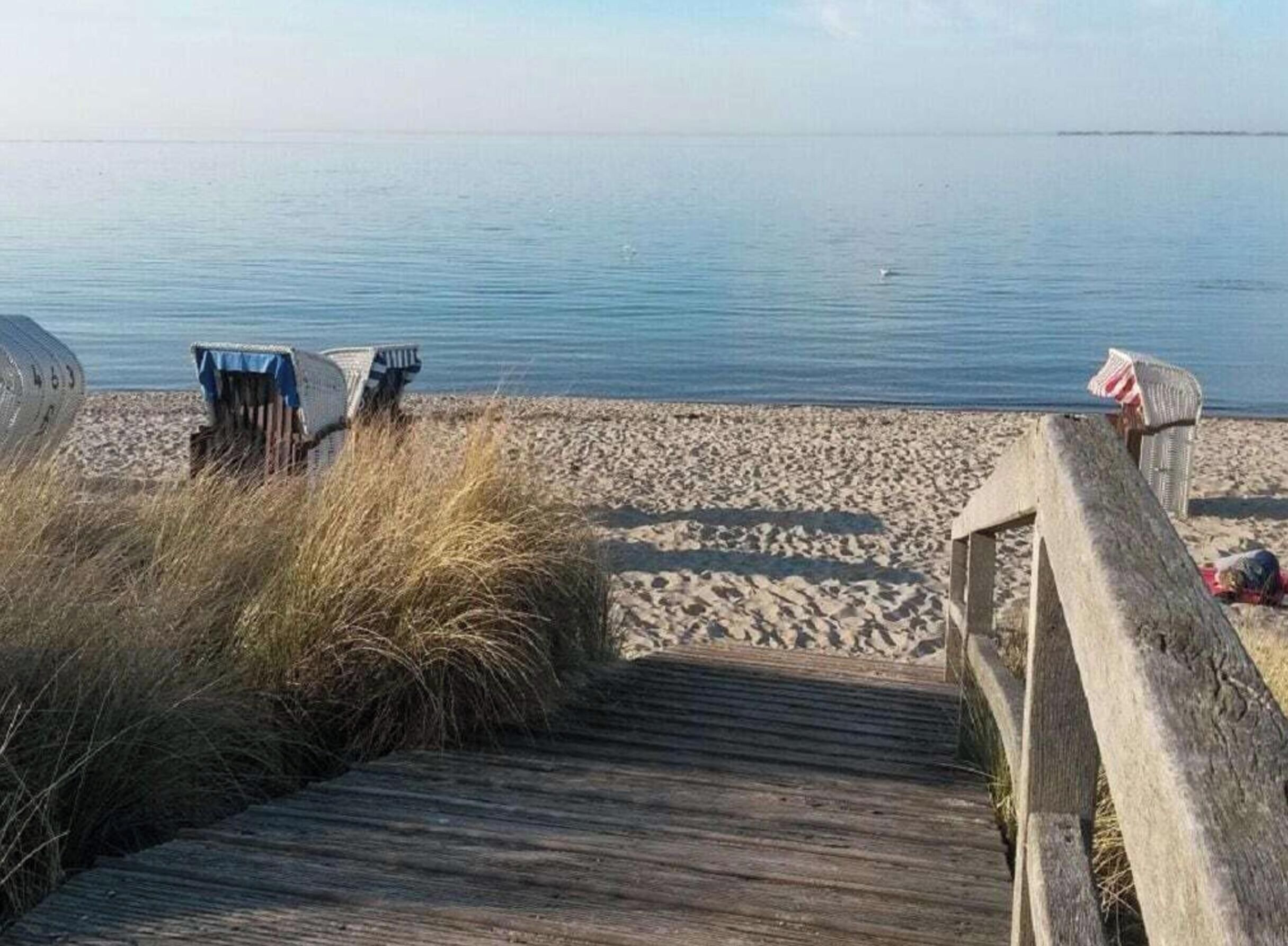 A wooden boardwalk leading to a sandy beach with beach chairs and a small structure.