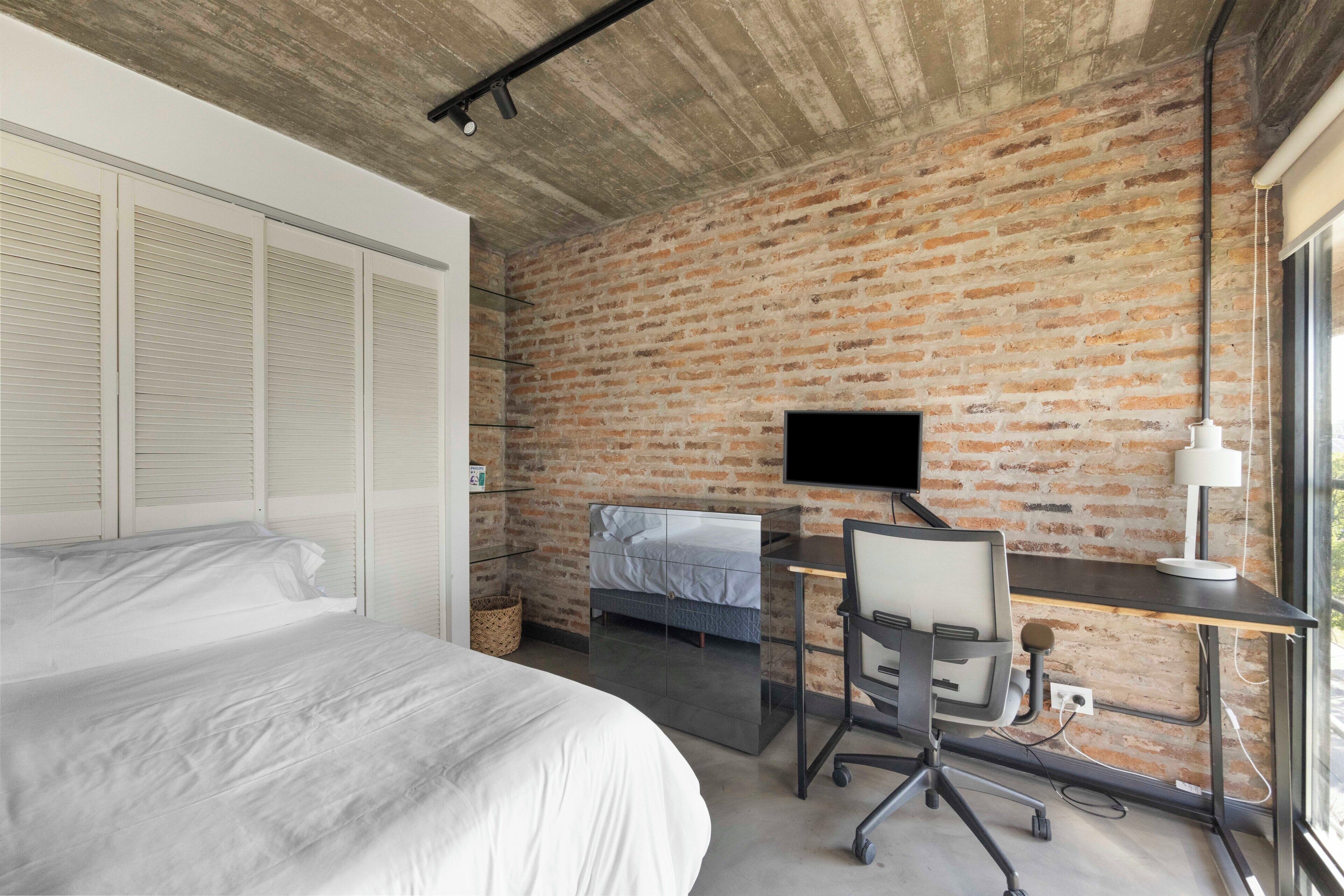 Un dormitorio con una cama grande, un escritorio con computadora, un televisor y una pared de ladrillo.