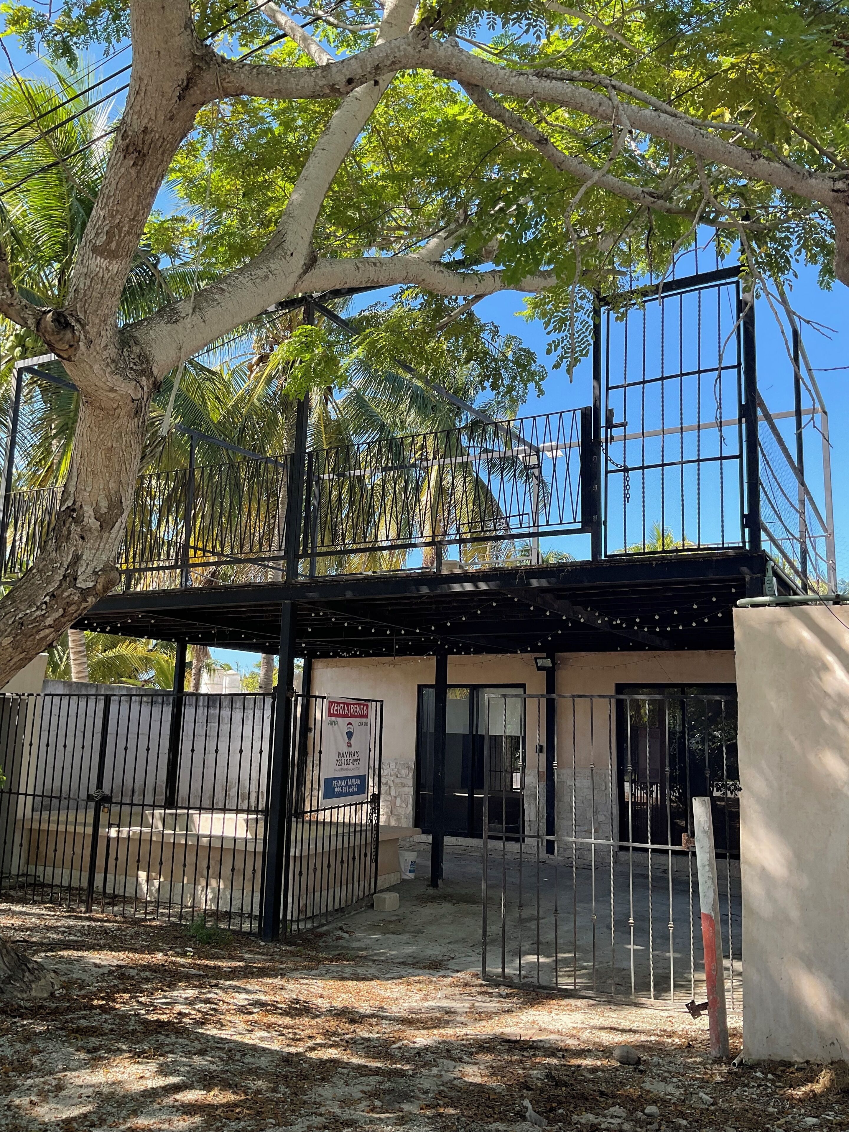 A gated property with a sign indicating rental availability, a covered entrance, and a tree providing shade.