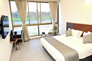 A hotel room with a bed, a desk, a chair, a TV, and a window with curtains. - Hotel Four Meadows (Mysuru)