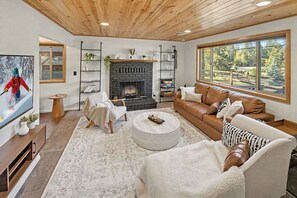A cozy living room with a fireplace, leather sofa, and a large window offering a view of the outdoors. - Big Bear Cabin - Hot Tub, Fire Pit, Pet-Friendly - 2 Min to Bear Mountain (Big Bear Lake)