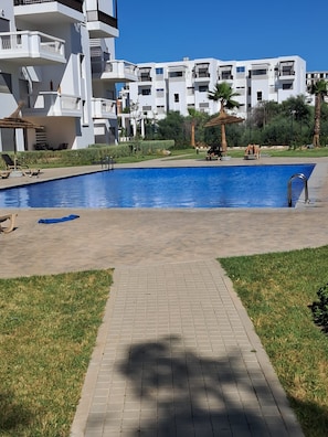 Ein Pool mit gepflastertem Gehweg, umgeben von Rasen und einem Gebäude mit Balkonen.
