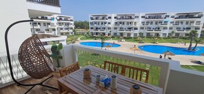 Ein Balkon mit einem Korbstuhl, einem Tisch mit Getränken und Blick auf einen Poolkomplex mit mehreren Gebäuden im Hintergrund.