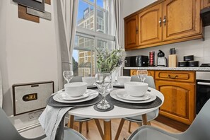 Ein Essbereich mit Tisch für zwei Personen, einer Blumenvase und Blick auf die Stadt durch große Fenster.
